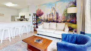 A living room with a white sofa, blue armchair, and a coffee table with remote controls. A mural of a castle is on the wall, and there is a kitchen area with white cabinets and a microwave. - Resort home with private pool, hot tub, tennis, game room, gym, sports courts (Kissimmee)