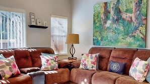 Una sala de estar con un sofá seccional marrón, almohadas coloridas, una l ámpara y un cuadro grande con motivos florales en la pared.