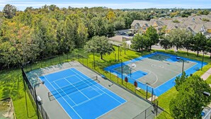Una cancha de tenis con cerco, rodeada de árboles y edificios residenciales.