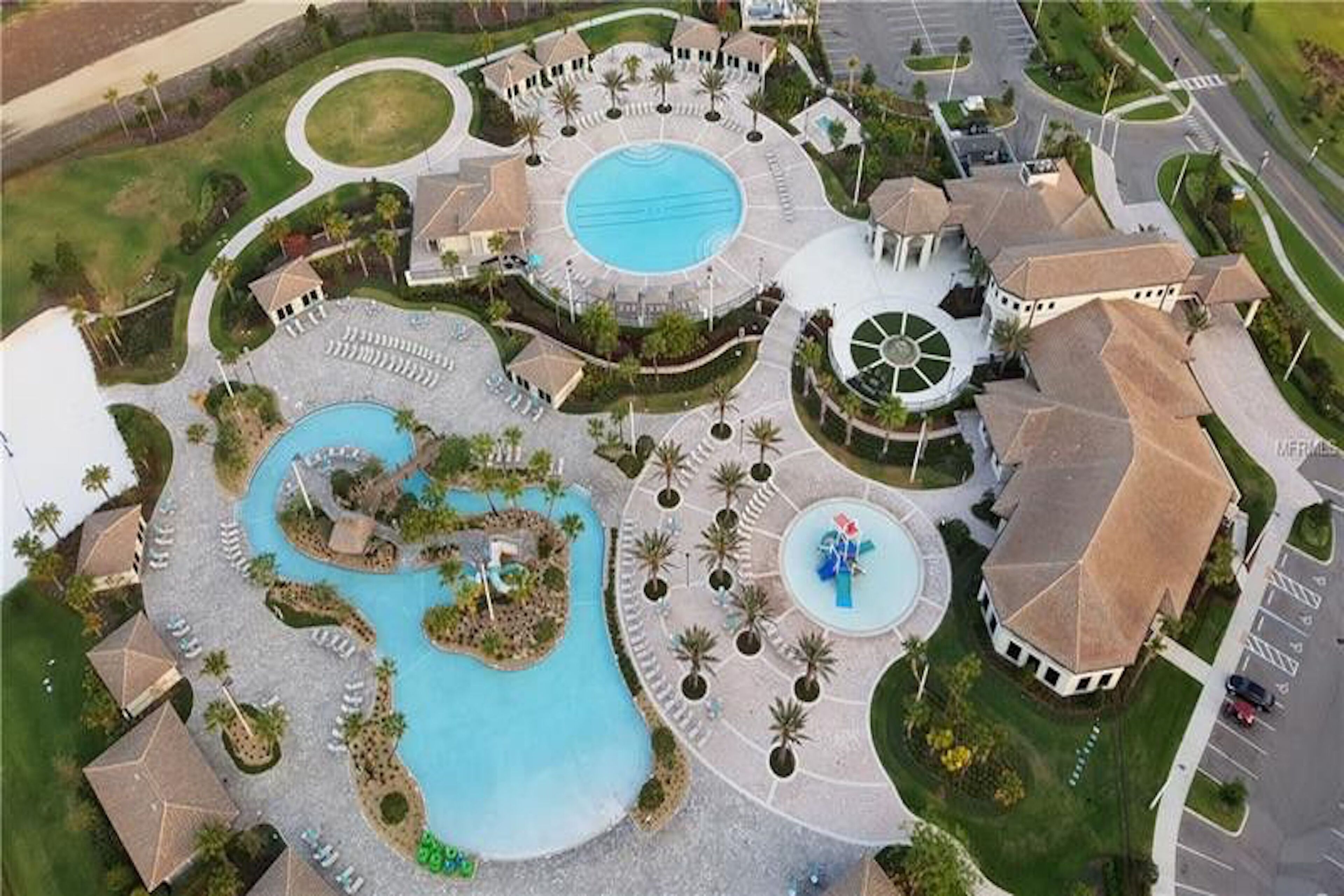 Vista aérea de un complejo turístico con una piscina central, varias edificaciones y zonas ajardinadas.