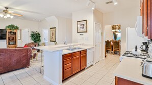Uma sala de estar espaçosa com um sofá marrom grande, uma área de jantar com cadeiras de madeira e uma cozinha com ilha central, armários brancos e uma geladeira.