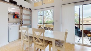 Una cuina moderna amb rentadora integrada, zona de menjador amb taula i cadires blanques, i grans finestres que donen a l'exterior.