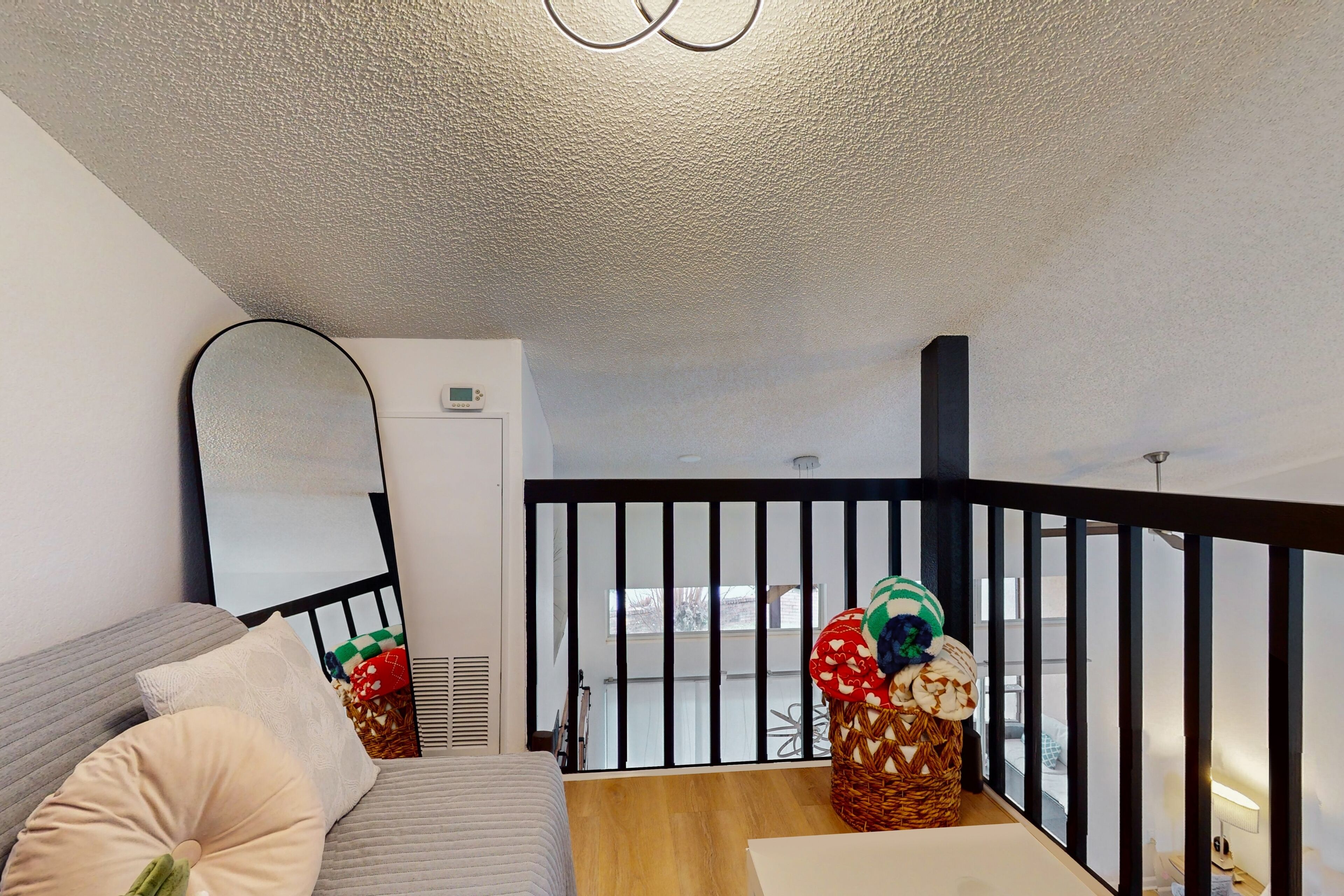 A room with a grey sofa, a large mirror, a wooden floor, a black railing, and a wicker basket with decorative items.