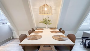 Uma sala de jantar com uma mesa comprida, seis cadeiras, jogos americanos trançados, uma planta central e um lustre.