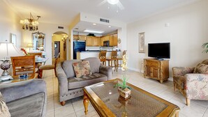 Uma sala de estar ampla com um sofá, poltronas, uma mesa de centro e uma televisão. Uma cozinha é visível ao fundo.
