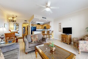 A spacious living room with a sofa, armchairs, a coffee table, and a television. A kitchen is visible in the background. - Welcoming lakefront condo with gorgeous lake view, kitchen, AC, pool & hot tub (Orlando)