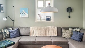 A modern living room with a grey sectional sofa, two round wooden side tables, a white fluffy throw blanket, a pendant light, a framed picture, a clock, and a window with blinds. - 2 bedroom stunning home in Haugesund (Haugesund)