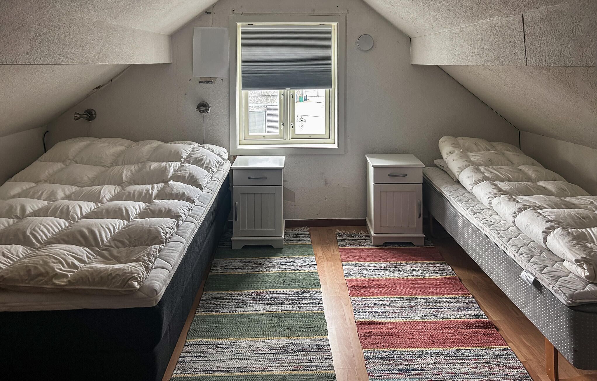 A small, triangular attic bedroom with two single beds, a window with blinds, a small white dresser, and a colorful striped rug.