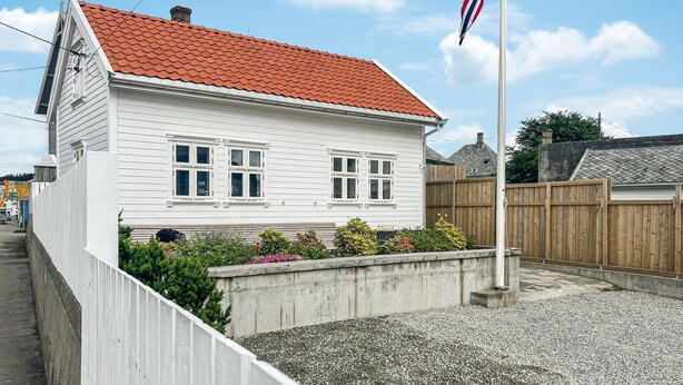Une maison blanche avec un toit recouvert de tuiles rouges, un mât sur lequel flotte un drapeau et une allée gravillonnée.