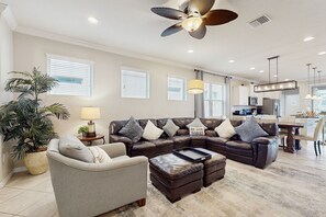 Ein geräumiges Wohnzimmer mit einem Ecksofa, einem grauen Sessel und einem Couchtisch. Es gibt einen Deckenventilator, eine Topfpflanze und eine Lampe.
