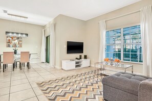 Una sala de estar moderna con un televisor de pantalla plana, un sofá, un comedor con sillas y un amplio ventanal con persianas.