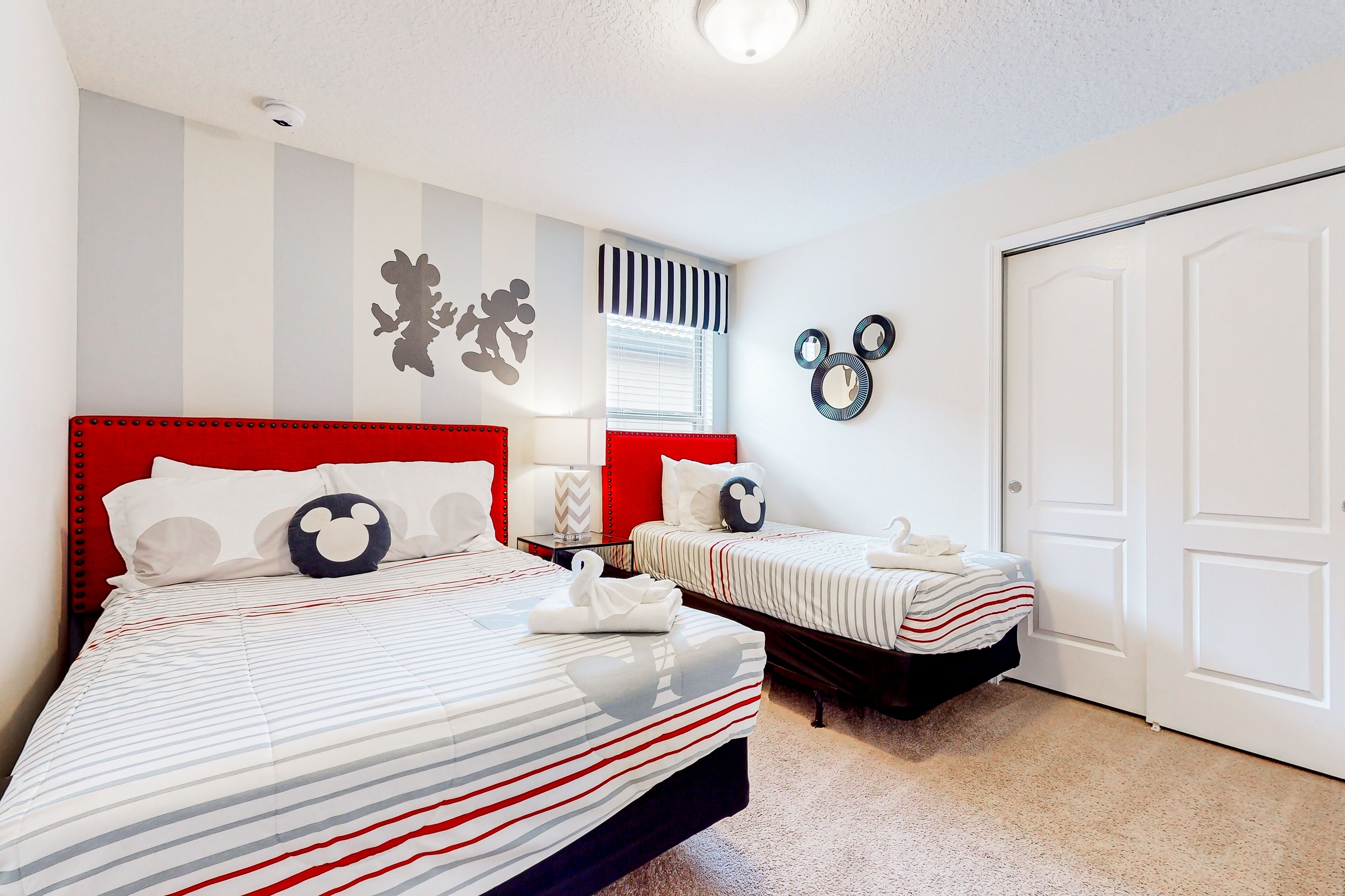 Un dormitorio con dos camas, ropa de cama a rayas, cabecera roja, almohadas de Mickey Mouse y decoraciones en la pared que incluyen siluetas de Mickey Mouse y espejos circulares.