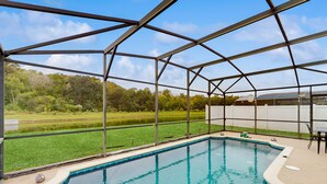 A swimming pool with a metal roof structure, surrounded by a grassy area and a view of trees. - Inviting pool home with tranquil pond view near theme parks & attractions (Kissimmee)