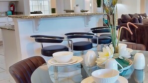 Een moderne eetruimte met een glazen tafel voor vier personen, omringd door donkere lederen stoelen. In het midden staat een decoratief middelpunt met witte bloemen en groene bladeren.