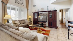 Amplia sala de estar con un juego de sofá, una mesa de centro de vidrio y un televisor. Se observa un aparador de madera con objetos decorativos y un espejo grande.