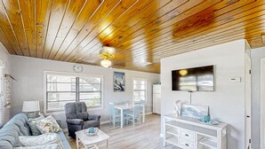 A cozy living room with a wooden ceiling, a sofa, a dining table with chairs, a TV mounted on the wall, and a small white desk. - Lake June Resort #9 | Condo in Lake Placid (Lake Placid)