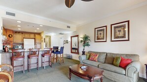 A spacious living room with a sofa, coffee table, and chairs. There is a kitchen area with a refrigerator and cabinets.