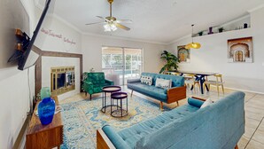 Ein geräumiges Wohnzimmer mit einem blauen Sofa, einem grünen Sessel und einem Couchtisch. Ein Deckenventilator und ein Fernseher sind an der Wand angebracht.
