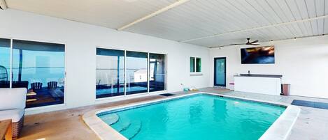 A modern indoor swimming pool with clear glass walls, a ceiling fan, and a view of the cityscape.