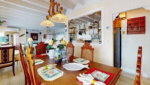 Ein Essbereich mit einem Holztisch, gedeckt für vier Personen, umgeben von Holzstühlen. Über dem Tisch hängt eine Chandelier, und im Hintergrund ist eine Küche zu sehen.