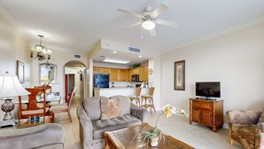 A living room with a sofa, coffee table, and TV, connected to a kitchen with wooden cabinets and a ceiling fan. - Inviting lakefront condo near attractions with lake view, AC, pool & hot tub (Orlando)
