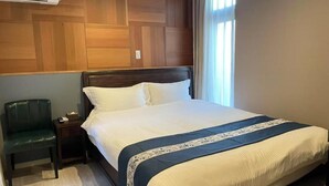 Ein ordentlich bezogenes Bett mit Holz-Kopfteil, ein Nachttisch mit Lampe und ein Stuhl.