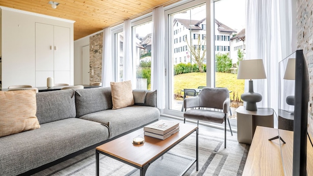 Ein modernes Wohnzimmer mit grauem Sofa, einem Holztisch und einem grauen Sessel. Ein großes Fenster bietet Blick auf ein Gebäude und Grünflächen.
