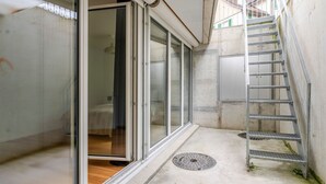 Ein schmaler, geschlossener Raum mit einer Schiebetür aus Glas, die zu einem Schlafzimmer führt, einer Betonwand und einer Metalltreppe.