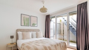 Ein Schlafzimmer mit einem Bett, einem Nachttisch, einer Lampe und einem großen Fenster mit Vorhängen.