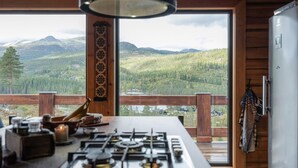A kitchen with a gas stove, wooden cabinets, and a view of a forested mountain range through large windows. - 4 bedroom amazing home in Tuddal (Tuddal)