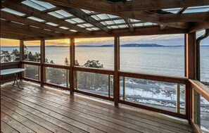 Una terraza de madera con ventanas amplias que dan a un cuerpo de agua y un paisaje cubierto de nieve.