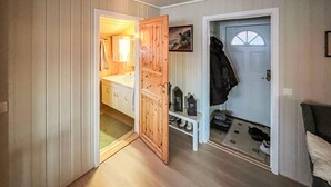 Ein Flur mit einer Holztür, die zu einem Badezimmer mit Waschbecken und Dusche führt. Ein Kleiderständer mit einem aufgehängten Jackett und ein Schuhregal mit Schuhen und einer Laterne stehen dort.
