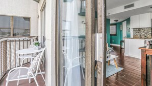 Ein Balkon mit einem weißen Tisch und Stühlen, eine Glastür, die zu einer Küche mit türkisfarbenen Wänden und Holzboden führt.