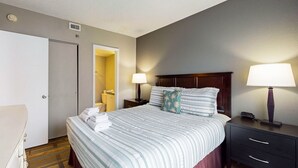 A hotel room with a bed, two bedside lamps, a nightstand, a wooden headboard, a door, and a bathroom visible through an open door. - Spacious Condo with a Kitchen, AC, Pools, Hot Tubs, Tennis, & Gym (Orlando)