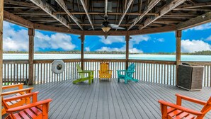 Una terraza techada con barandillas de madera, un ventilador de techo y tres sillas Adirondack de colores frente a una masa de agua.