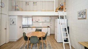 Een compacte keuken met een slaapvide erboven, met een witte balustrade, een houten eettafel en een ladder naar de slaapvide.