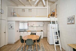 A compact kitchen with a loft bedroom above, featuring a white railing, a wooden dining table, and a ladder to the loft. - Beachfront 1BR + Loft. Surf-Café-Unwind (Sunshine Coast)