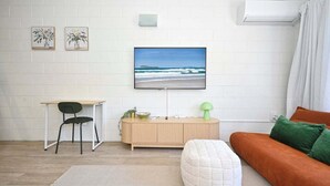 Een woonkamer met een aan de muur bevestigde televisie, een houten dressoir, een groene lamp, een bruine bank en een witte poef.