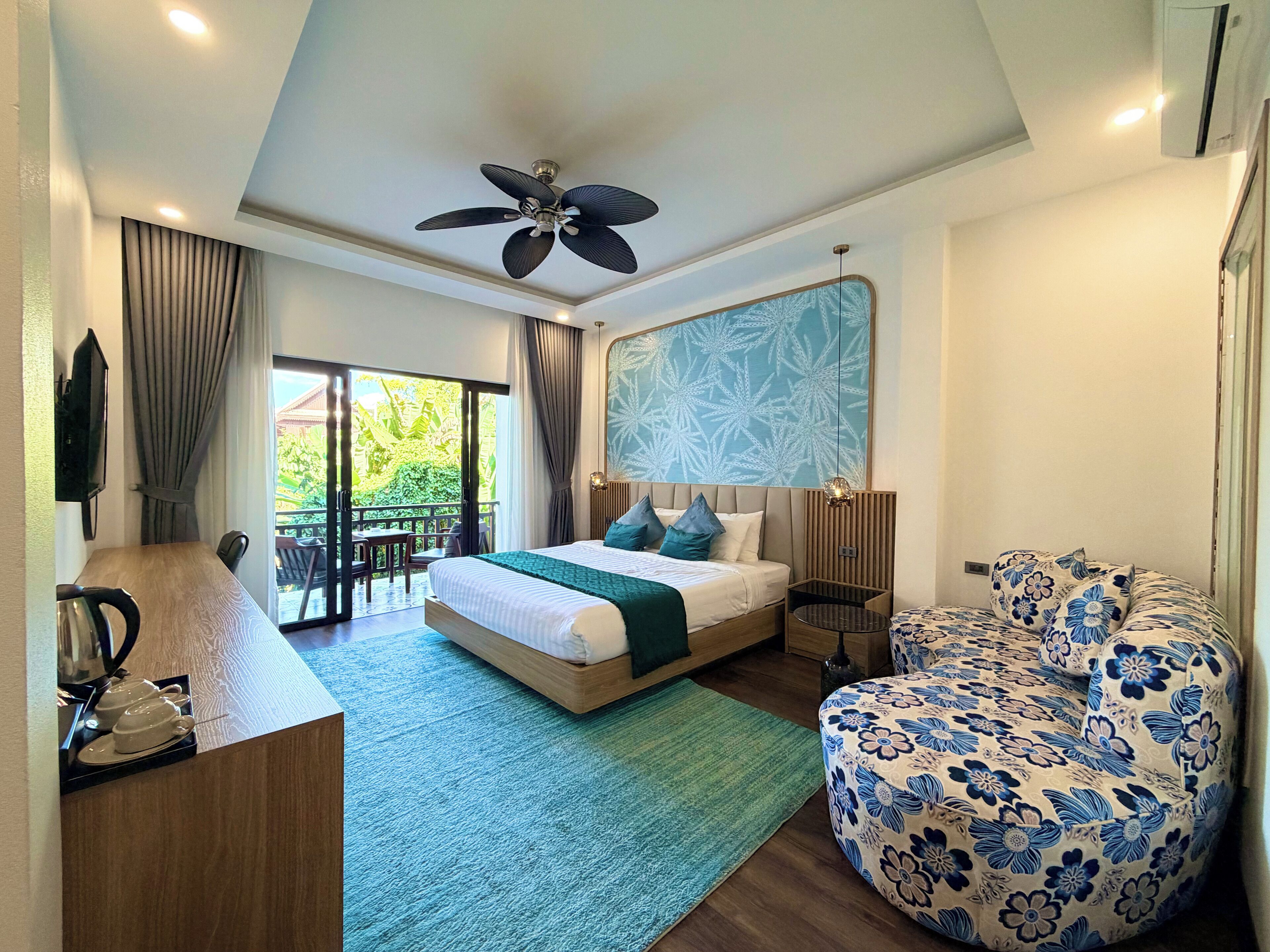 A hotel room with a bed, a desk with a kettle, a chair, a ceiling fan, and a large window with curtains.