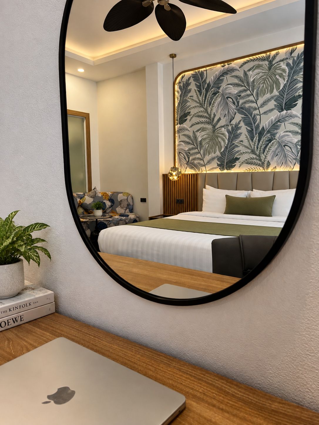 Une chambre à coucher avec un lit, un bureau avec un ordinateur portable, une plante en pot et un mur orné d’une fresque à motifs de feuilles.