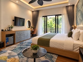 A modern hotel room with a bed, a desk, a television, and a balcony with a view of greenery.