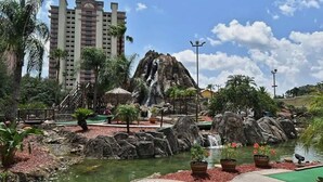 A resort area with a waterfall, palm trees, and a large building in the background. - Lakefront condo with beautiful views, wifi, central AC, shared pool, & hot tub (Orlando)