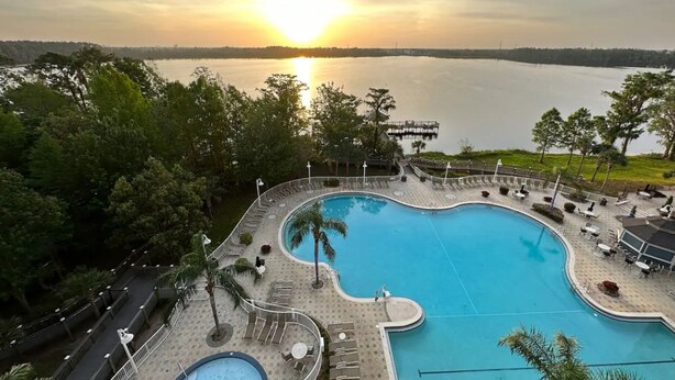 A swimming pool with a curved edge, surrounded by lounge chairs and palm trees, with a view of a lake and sunset in the background. - Lakefront condo with beautiful views, wifi, central AC, shared pool, & hot tub (Orlando)