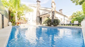 Una piscina con bordo a mosaico geometrico, circondata da vegetazione rigogliosa e una casa moderna con un camino ben visibile.