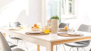 Una tavola imbandita per la colazione, con tovaglia bianca, sottopiatti intrecciati, una ciotola di frutta fresca, un bicchiere di succo d'arancia e una pianta in vaso.