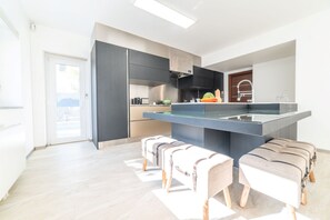 A modern kitchen with dark cabinetry, a central island, and a large window. - Villa Solmaris | Costa da Caparica | Private Pool - Jacuzzi | Near Beach (Lisbon)