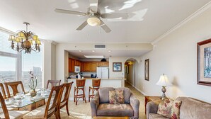 Ein geräumiges Wohnzimmer mit einem Esstisch mit Glasplatte, Holzstühlen, einer Couch und einem Couchtisch. Über dem Essbereich sind ein Deckenventilator und eine Chandelier zu sehen.