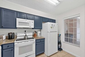 A kitchen with white appliances, blue cabinets, and a window with blinds. - 2BR Worldquest Retreat in Orlando near Disney (Orlando)