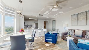 Una sala moderna con comedor, una cocina al fondo y ventanas amplias con vista a la ciudad.