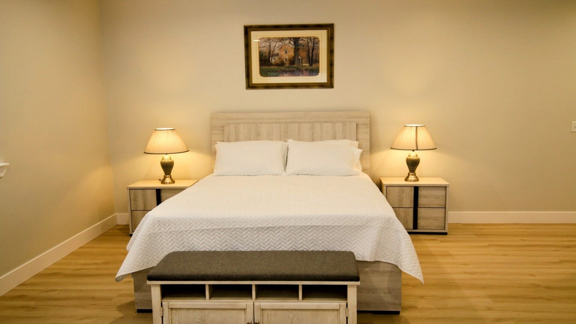 A neatly made bed with a white quilt, flanked by two bedside tables with lamps, a bench at the foot of the bed, and a framed picture on the wall.